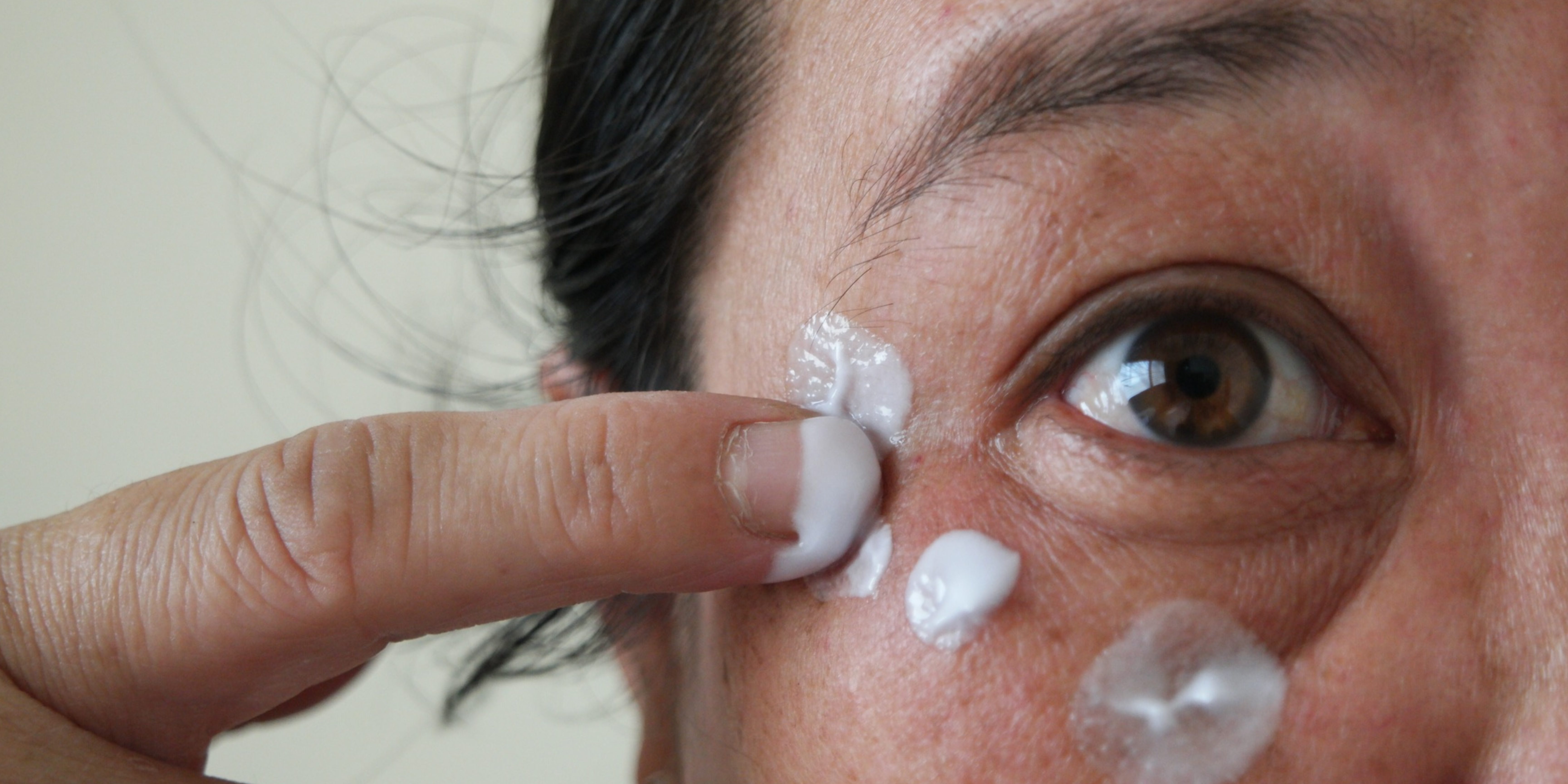 A closeup picture of a woman's eye. She is applying cream to the area around her eye her e