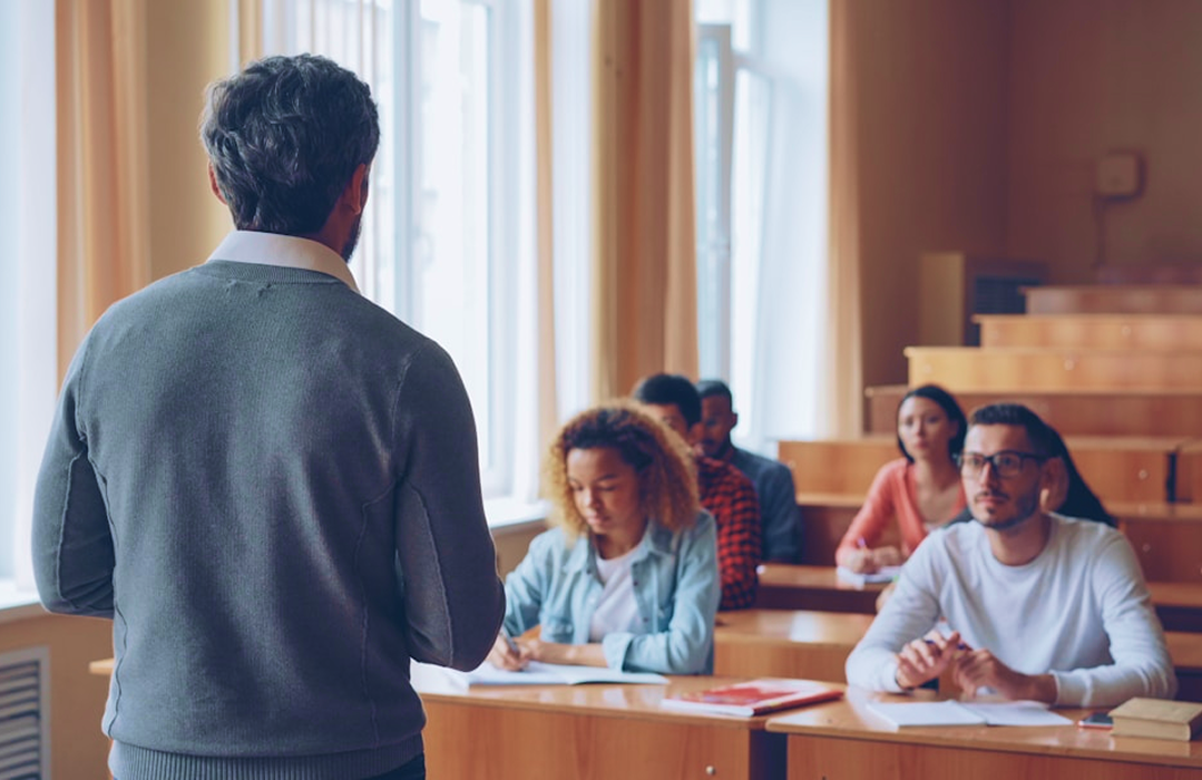A man with their back to camera speaks to a group of people sat down within a classroom