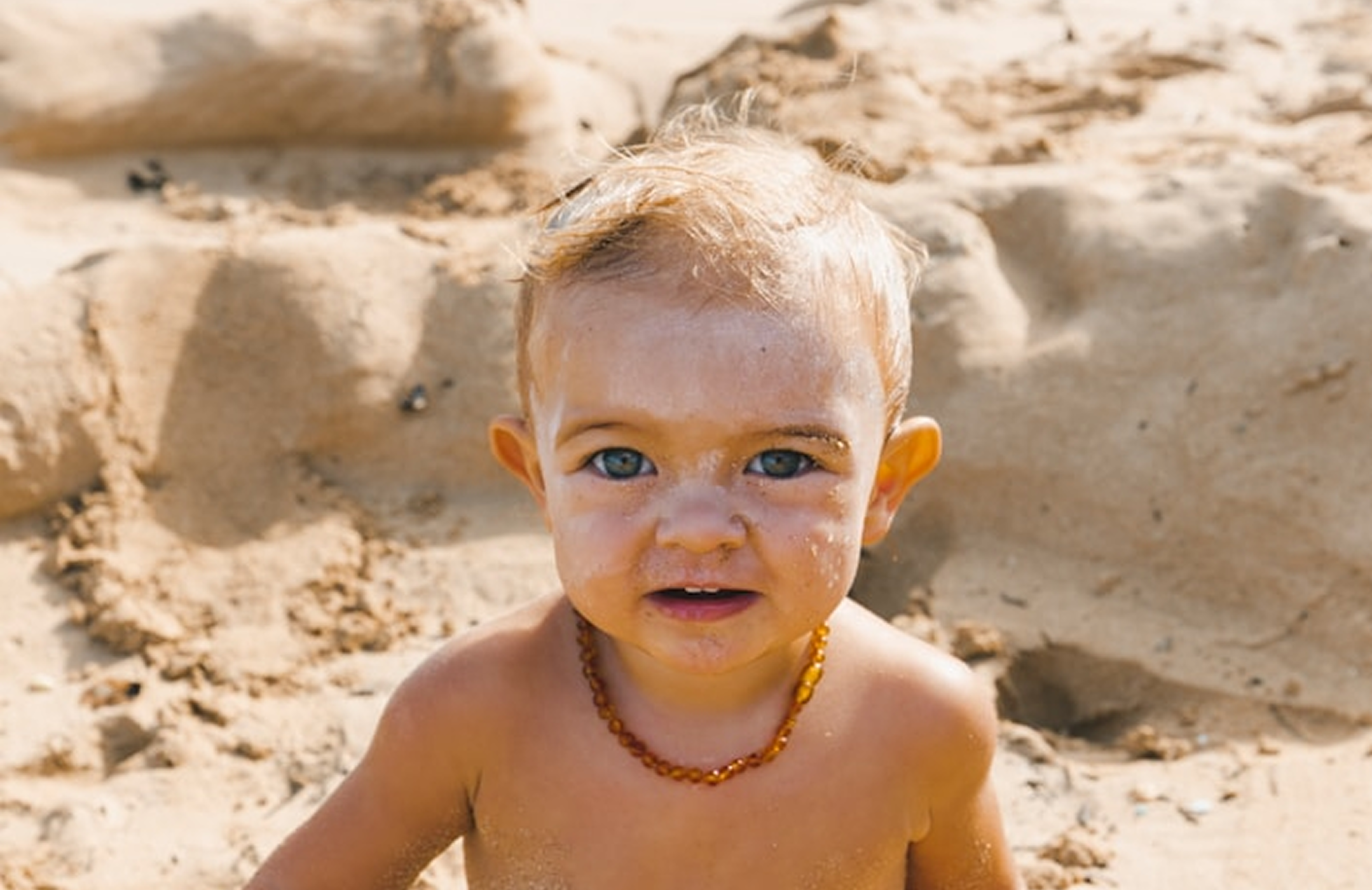 A young child is on the beach facing the camera wearing a necklace and covered in sand and sun cream